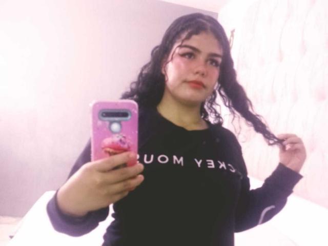 BrianaCleython with curly black hair, wearing a black long-sleeve shirt, holding a pink phone with a red pop socket, taking a selfie with a slight smirk.
