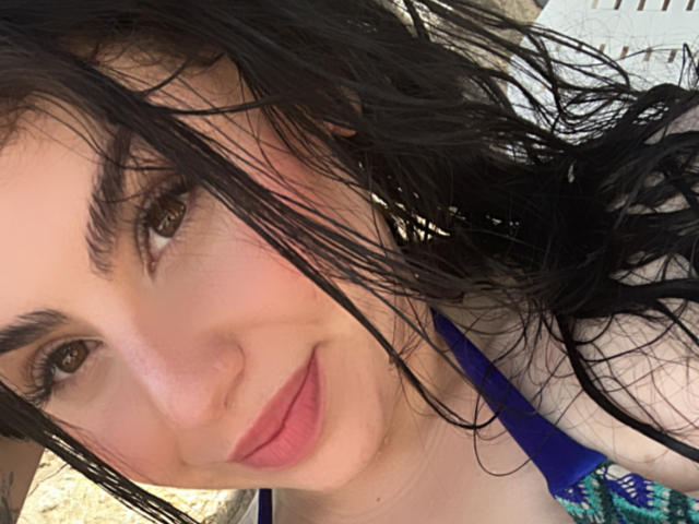 VenusxMusa with wet black hair, brown eyes, and pink lips, wearing a blue top, smiling slightly at the camera. Her eyebrows are thick and she has a small tattoo.