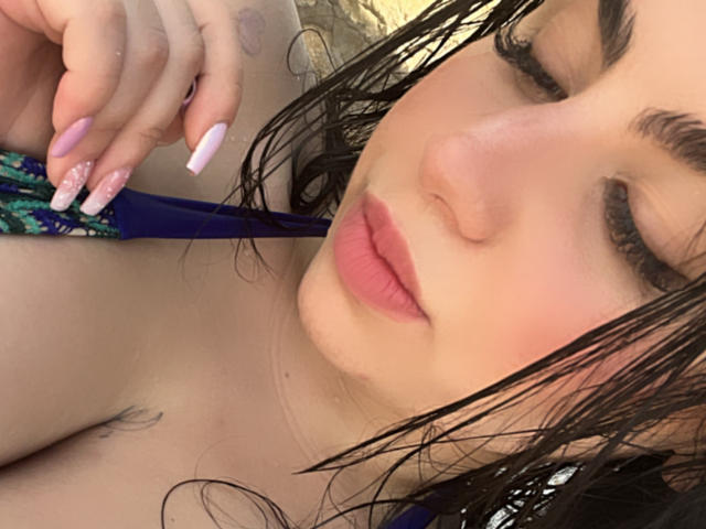 VenusxMusa with wet black hair, pink lips, and pink nails, holding a blue strap, showing a tattoo on her shoulder. Her eyes are half-closed, and she's looking down.