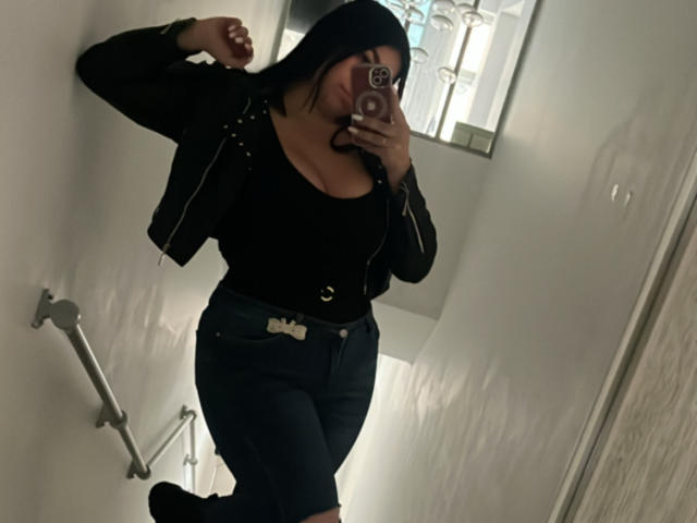 VenusxMusa with black hair, black jacket, black top, and dark jeans, holding phone, taking mirror selfie, showing her ass in tight jeans, in dimly lit bathroom.