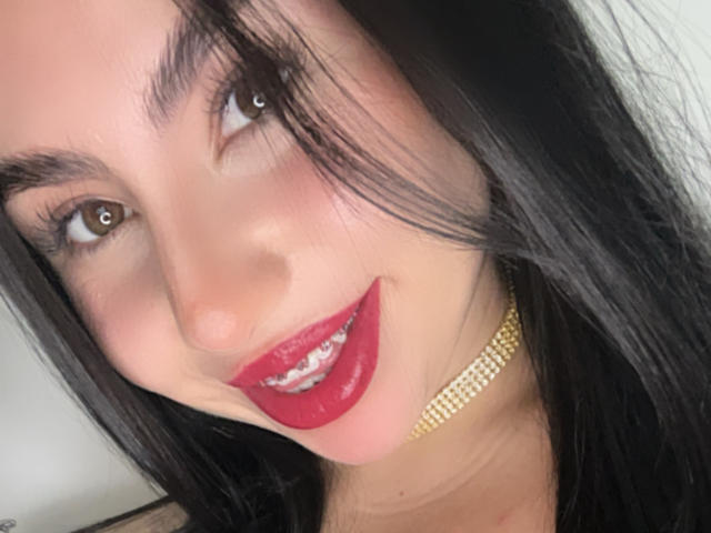 VenusxMusa has dark black hair, brown eyes, red lipstick, and a gold choker. She's smiling with visible teeth. Her face is slightly tilted to the left.