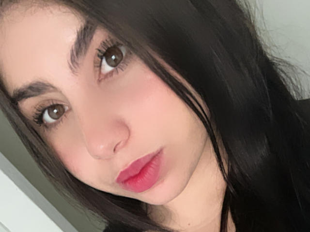 VenusxMusa has brown eyes, dark eyebrows, long black hair, and pink lipstick. Her face is slightly angled, showing a neutral expression. She's wearing a black top.