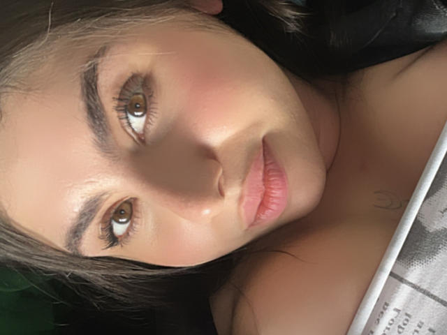 VenusxMusa's face with brown eyes, pink lips, and slight blush, lying down, showing bare shoulders, dark brown hair, and subtle tattoo on chest. Soft, natural light.