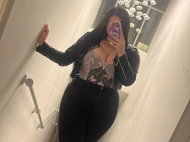 VenusxMusa with big tits, wearing a black leather jacket and floral bra, black pants, standing in a white-walled room, taking a mirror selfie with a purple phone case.