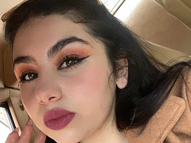VenusxMusa has pink lips, brown eyes with black eyeliner, orange eyeshadow, and dark eyebrows. She's wearing a brown hoodie, with long black hair and tan earbuds.