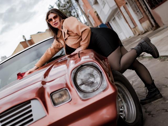 VenusxMusa è appoggiata a una macchina rossa, indossa una giacca color sabbia, una gonna nera e stivali neri, con i glutei in evidenza e gli occhiali da sole, mostrando il seno e le calze..