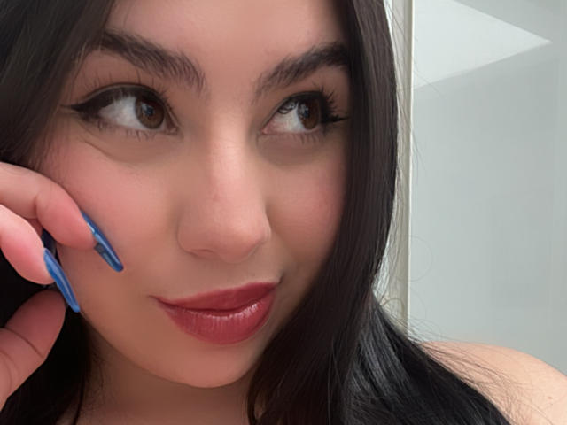 VenusxMusa, con lunghi capelli neri, occhi marroni e rossetto rosso, tiene un eyeliner blu vicino alla guancia, sorridendo leggermente. Le sue unghie sono dipinte di blu..