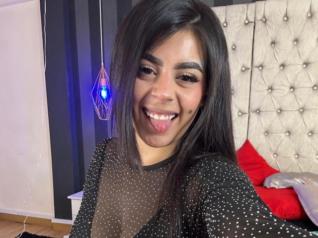 CamilePretty with dark hair, black sheer top with white polka dots, sticking out tongue, smiling, blue hanging light, red and silver pillows, beige tufted headboard.