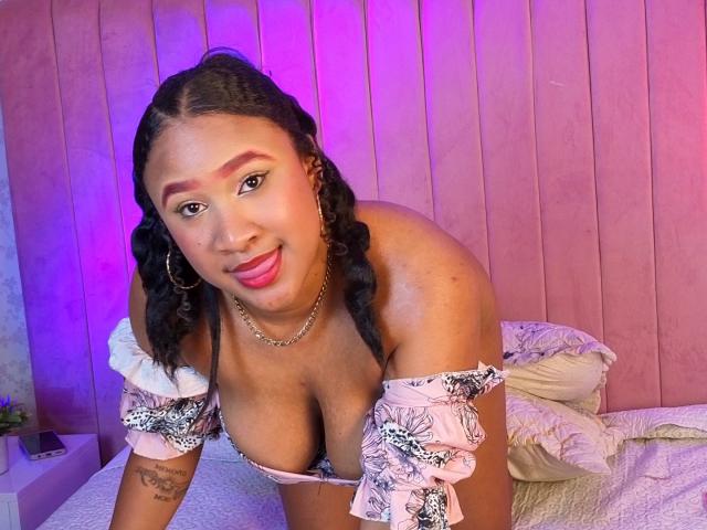MikaelaCooper with braided hair, pink off-shoulder top, showing big tits, smiling, purple and pink lighting, hoop earrings, chain necklace, tattoo on left arm, leaning on bed.
