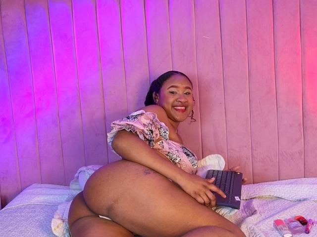 MikaelaCooper with dark skin, smiling, wearing floral top, big ass, laptop on bed, pink and purple lights, braided hair, red lipstick, white bedsheet, and pink nail polish.