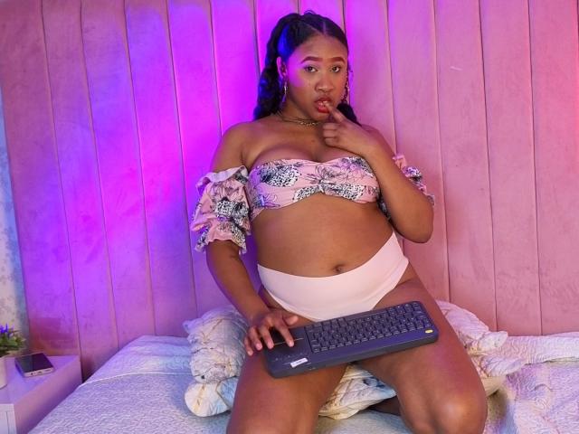 MikaelaCooper with brown skin, curly hair, pink floral off-shoulder top, white panties, holding keyboard, finger in mouth, purple and pink lighting, sitting on bed with pillows.