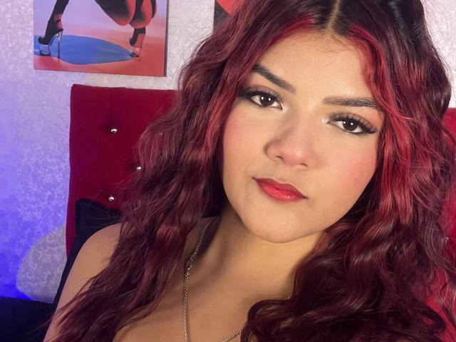 VelvetFlame has red wavy hair, dark eyebrows, long eyelashes, red lipstick, and a slight smile. She's wearing a necklace and a red headboard is behind her.