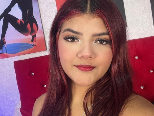 VelvetFlame with red-dyed hair, full eyebrows, long eyelashes, and red lipstick, smiling at the camera, sitting against a red tufted headboard, with a sexy black-and-red poster.