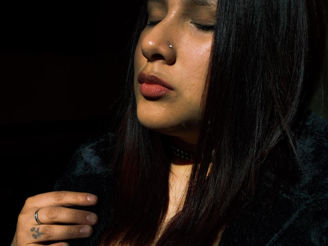 LadyStarOne has dark straight hair, a nose piercing, and closed eyes, wearing a black choker and fur, with a hand showing a ring and tattoo, lit by sunlight.