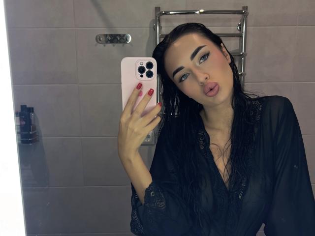 Adeliss with wet black hair, pouty lips, and blue eyes, wearing a sheer black robe, taking a mirror selfie with pink phone, red nails, in a bathroom.