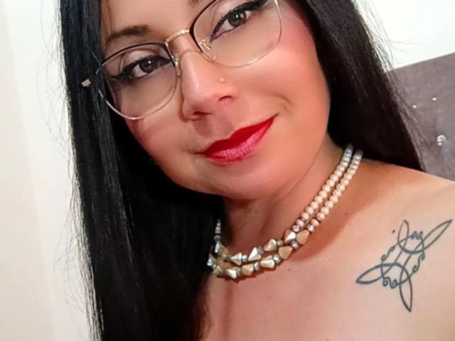 Zaria69 has long black hair, wears glasses, red lipstick, and a pearl necklace, smiling with a tattoo on her right shoulder. Her tits are slightly visible.