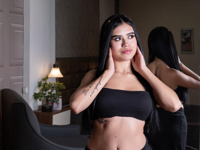 Danielle Harper, met lang zwart haar, draagt een zwarte top zonder bandjes, waarbij tatoeages op haar armen zichtbaar zijn. Haar handen zijn op haar gezicht, het is een spiegelbeeld, en ze heeft een zelfverzekerde glimlach. De scène speelt zich af in een slaapkamer..