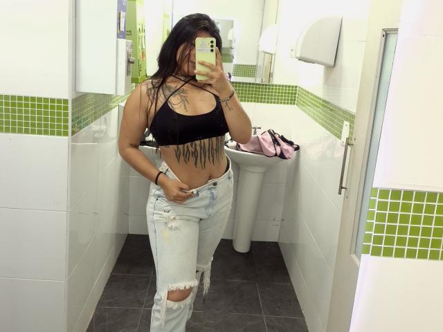 DesireSweet with wet black hair, wearing a black sports bra showing her tits, and ripped jeans, has a tattooed belly and is taking a mirror selfie in a bathroom.