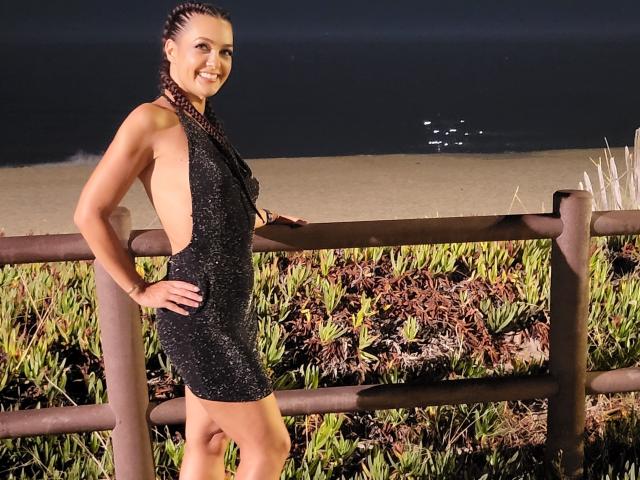 SelinaKylla stands in a black, side-open, sparkly dress, showing her ass and back, with braided hair, smiling, hands on a wooden fence, by a dark ocean.
