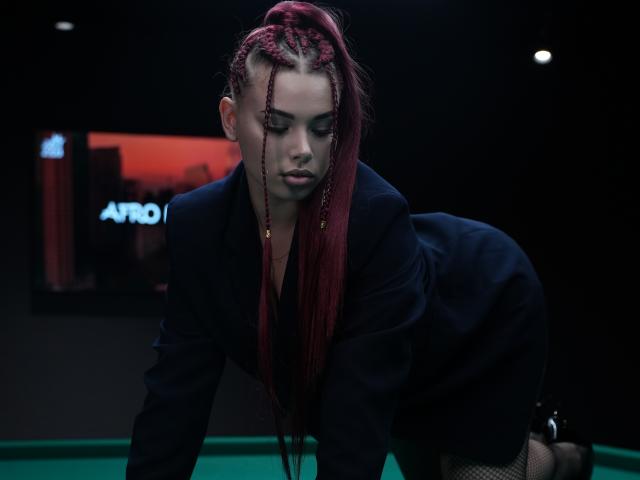 Niferttiti has red braids, a dark blazer, and is on all fours on a green pool table, looking down with a serious expression. Her ass is prominent.