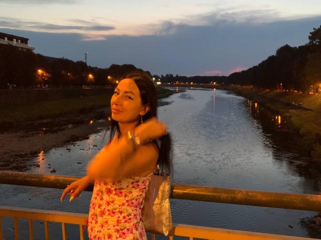 MirandaHott69 with long dark hair, wearing a pink floral dress and furry arm warmers, smiling at a river during dusk, standing by a yellow railing, holding a silver bag.