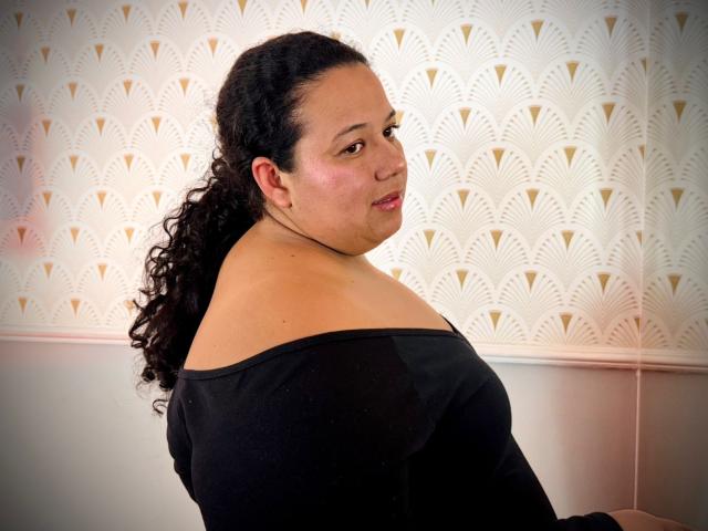 VioletthaBartomeu, curvy, with dark curly hair in a ponytail, black off-shoulder top, looking over her shoulder, smiling slightly, light skin, small nose, pink lips.