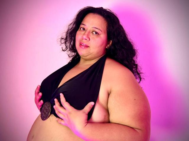 VioletthaBartomeu, plus-sized with curly black hair, holding black halter top to cover her big tits, pink and white light, slightly smiling, standing against plain background.
