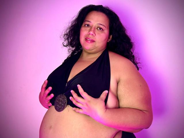VioletthaBartomeu, a plus-sized woman with curly black hair, holds her big tits with black hands, wearing a black halter top, and a large metal pendant, under pink lighting.