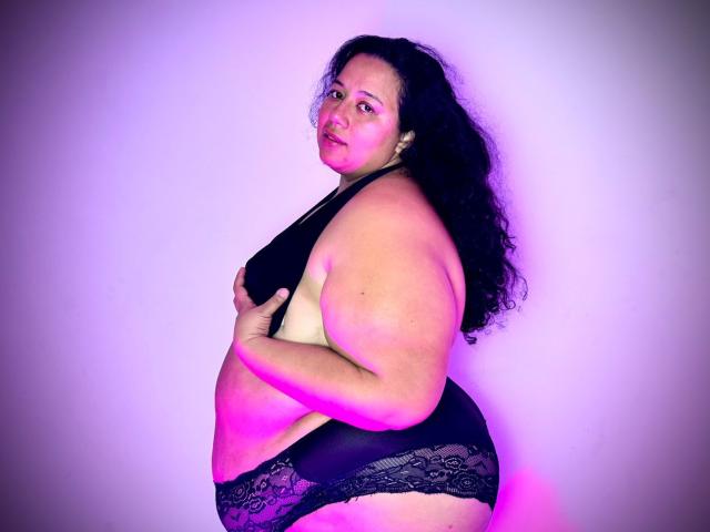 VioletthaBartomeu, plus-sized with curly black hair, wearing black lace underwear and a black bra, covering her tits with one hand, standing in a purple-lit background.
