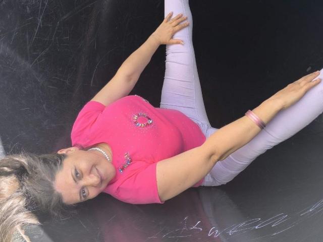 BlissSophia lying on her back, pink top, white pants, arms spread, legs up, smiling, pink bracelet, pearl necklace, blonde hair, black surface, visible scratches.