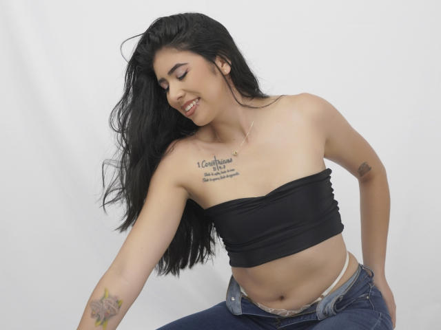 MiluSky, con cabello negro largo, sonriendo, vestida con un top negro ajustado, mostrando pequeños senos, tatuajes en el pecho y el brazo, jeans desabrochados, ropa interior blanca y un collar de plata..