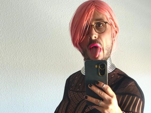 GingerPink-hot with pink hair, glasses, and black mesh top, tongue out, holding phone, showing ring, and painted black nails, looking in mirror.