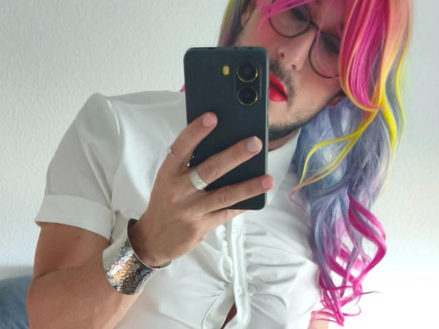 GingerPink-hot with rainbow-colored hair, red lipstick, glasses, white shirt, and silver bracelet, taking a selfie with a black phone, wearing a ring on their right hand.