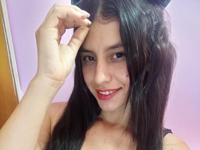 MadissonFontaine with long black hair, smiling with red lipstick, hand touching her head, showing her cute face and small moles. Her boobs are slightly visible in a black top.