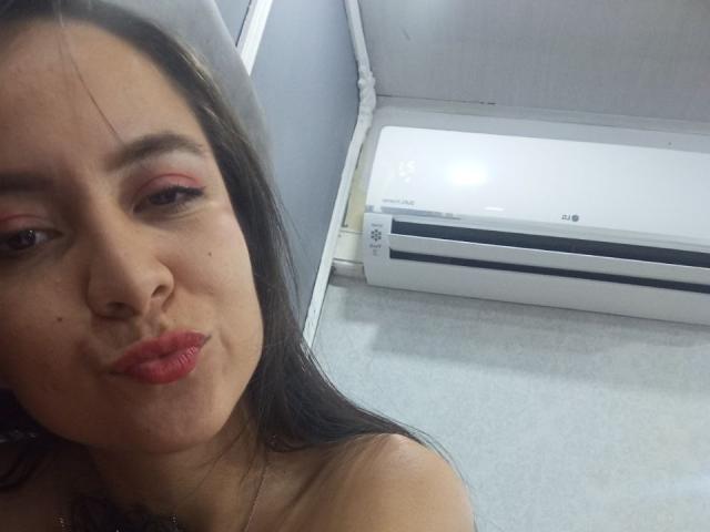 JessyLenon with red lipstick, pouty lips, and long brown hair, looking at camera, next to a white LG air conditioner. Boobs not visible, focus on face.