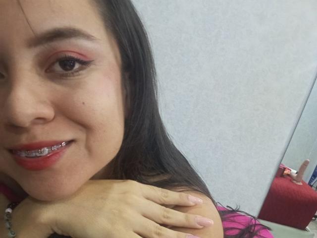 JessyLenon with red eyeshadow, pink lipstick, and metal braces, smiling, dark hair, holding shoulder, pale skin, right eye visible, pink nail polish, white background, red chair reflection.