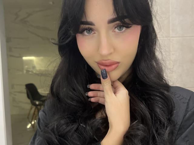 Adeliss, com cabelos pretos longos, olhos azuis, blush rosa e batom escuro, tocando seus lábios rosados com o dedo, unhas pretas, vestindo uma blusa preta, olhando para a câmera..