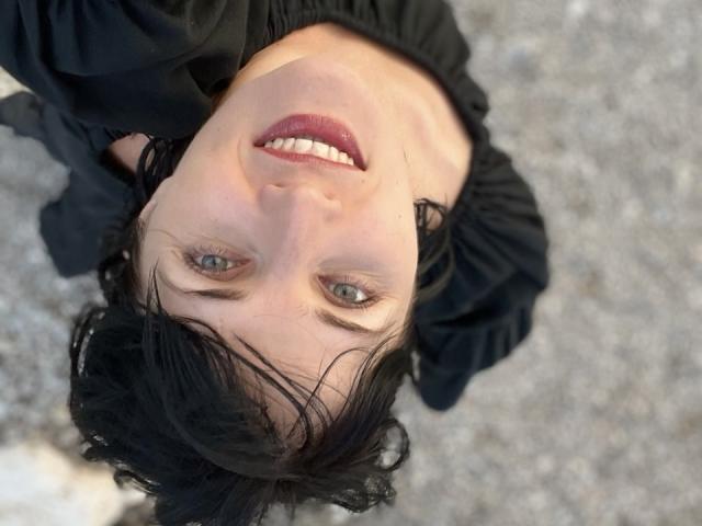 Le visage inversé d'AngelaFortune, avec ses cheveux noirs, son rouge à lèvres rouge et ses yeux gris, portant une robe noire, regardant vers le haut avec un sourire. Ses seins ne sont pas visibles..