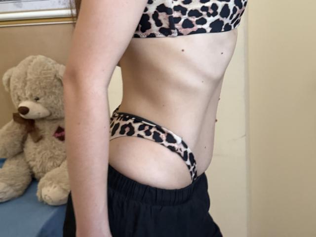 Stefanni's side profile showing a slim, pale body with small tits and a tiny, leopard-print ass in matching bra and panties, wearing black pants. A beige teddy bear is in the background.