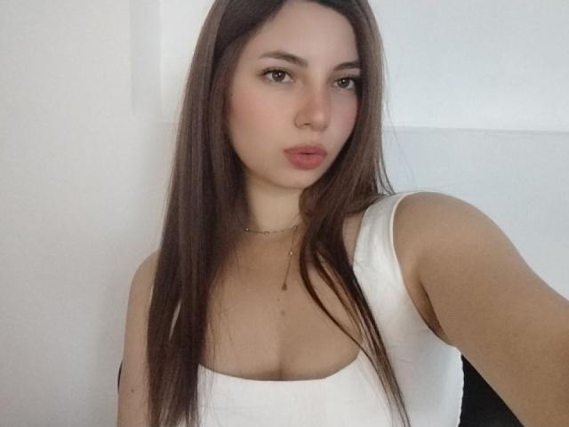 LinaFoxyHot has long brown hair, wearing a white tank top that shows her small boobs, with a neutral expression and slightly pouted lips, against a plain white background.