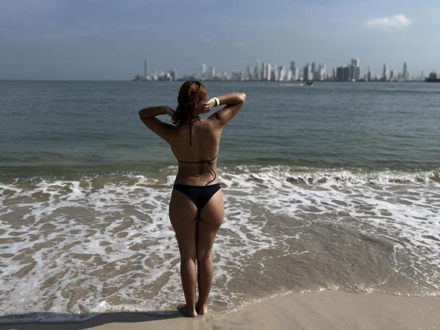 AlyMartiny stands on a beach, facing the ocean, wearing a black bikini, showing her ass, back, and wet hair, with her hands behind her head, city skyline in background.