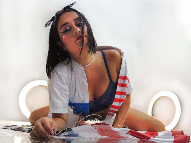 LunaSwt with dark hair, wearing a blue bra, white shirt with American flag, and necklace, leaning on a table with a flag, has hoop earrings and a small heart sticker.