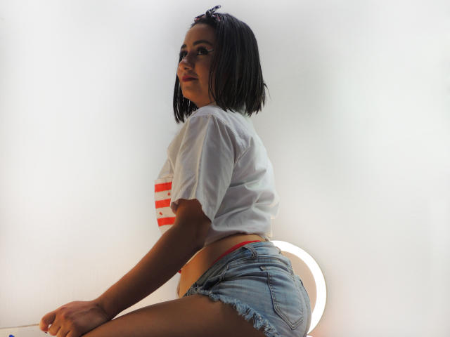 LunaSwt with a short black bob, white tee, and frayed denim shorts, sitting on a chair, showing her butt, and looking over her shoulder.