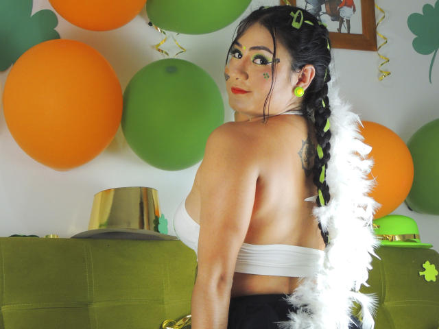 LunaSwt with green eye makeup, white strapless top, black skirt, white feather boa, braided black hair, yellow earrings, looking back, orange and green balloons, tattoo on right shoulder.