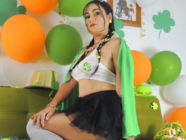 LunaSwt with green eye makeup, braided hair, white crop top with green shamrock, green jacket, black skirt, white knee socks, sitting, orange and green balloons, green wall.