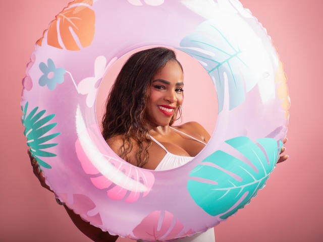 IsabellaKaned with curly brown hair, wearing a white top, holding a pink inflatable ring with tropical patterns, smiling with red lipstick, showcasing her tits through the ring.