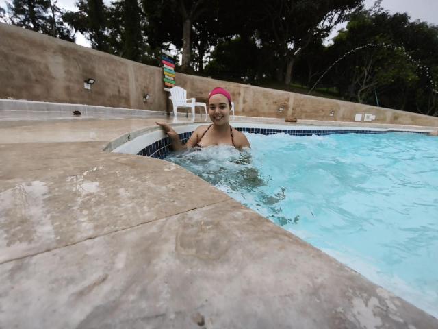 ZoeTobon69, pink swim cap, smiling, in a blue-tiled pool, partially submerged, concrete edge, white chair in background, trees, overcast sky, water splashing, medium skin tone, small tits.