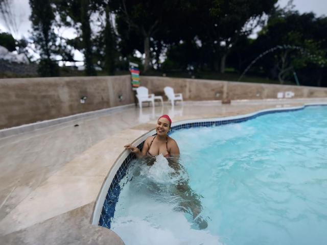 ZoeTobon69, dark-skinned with a red headscarf, sits in a blue-tiled hot tub, water splashing around her bare, medium-sized tits, smiling with one arm on the edge.