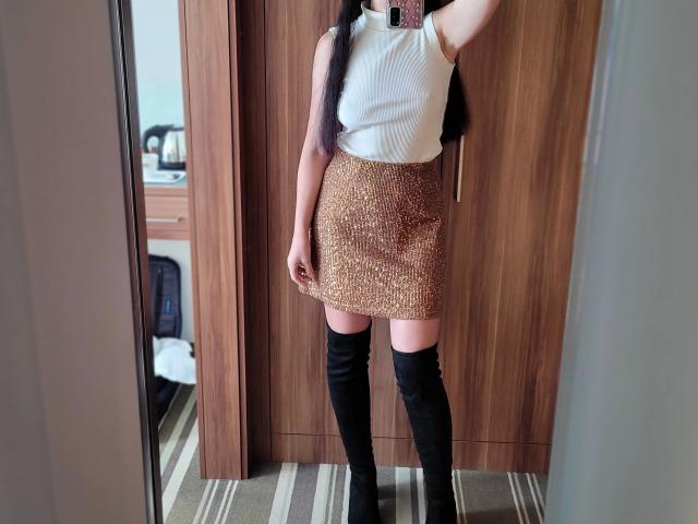 SweetJolina-hot wearing a white sleeveless top, gold glitter skirt, and black thigh-high boots, standing in front of a wooden door. Long black hair, pink phone case visible.