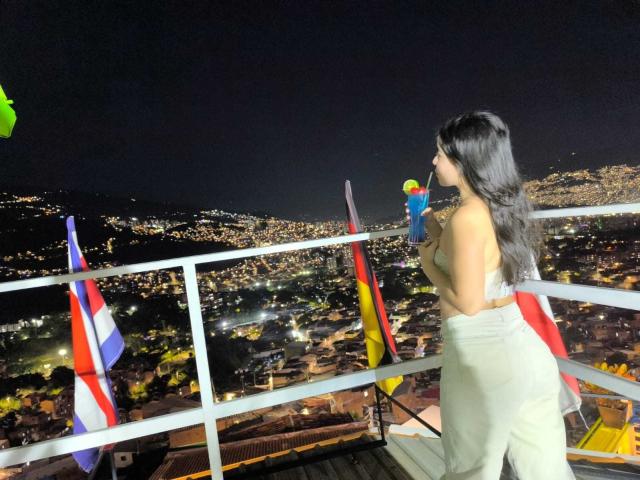 LissyCarter stands on a rooftop, topless with long black hair, holding a drink with a straw, wearing beige pants, and looking at a city illuminated at night.